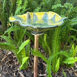 May include: A ceramic bird bath with a yellow and green leaf design. The bird bath is supported by a copper-colored pole and is surrounded by green ferns. The bird bath is designed to be placed outdoors.