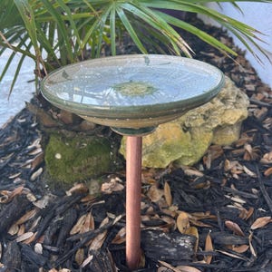 May include: A handcrafted ceramic bird bath with a green and beige glazed finish. The bird bath is mounted on a copper-colored metal pole. The bird bath is outdoors, surrounded by mulch, rocks, and greenery.