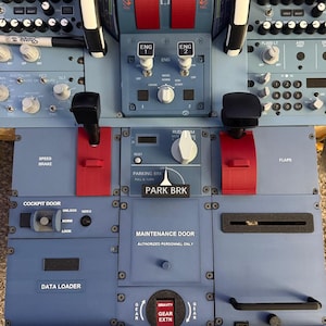 May include: A blue aircraft cockpit center pedestal featuring red throttle levers, a parking brake lever labeled PARK BRK, a gear extension lever, and various control panels with white text including SPEED BRAKE, FLAPS, and MAINTENANCE DOOR.