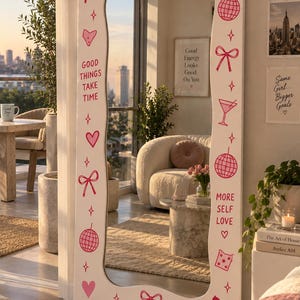 May include: A tall, rectangular floor mirror with a white frame featuring pink graphic designs including cherries, bows, hearts, disco balls, and stars. The text reads: You look good, GOOD THINGS TAKE TIME, and MORE SELF LOVE.