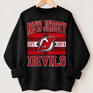 May include: A black crewneck sweatshirt featuring a red and white graphic print. The text reads NEW JERSEY DEVILS with EST. 1974. A central red diamond shape contains a stylised red and white NJ logo. The design has a distressed, vintage texture.