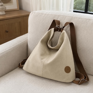Puede incluir: Una mochila de lona beige con silueta triangular, que cuenta con correas ajustables marrones y un parche circular de aspecto de cuero marr&oacute;n en la esquina inferior derecha. El bolso tiene una costura vertical central y herrajes met&aacute;licos.