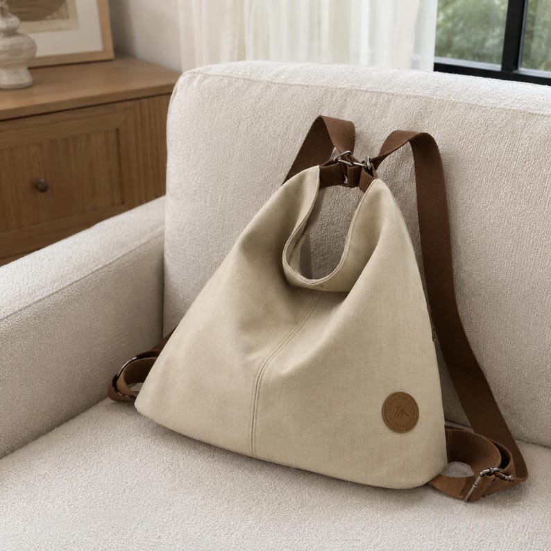Puede incluir: Una mochila de lona beige con silueta triangular, que cuenta con correas ajustables marrones y un parche circular de aspecto de cuero marr&oacute;n en la esquina inferior derecha. El bolso tiene una costura vertical central y herrajes met&aacute;licos.