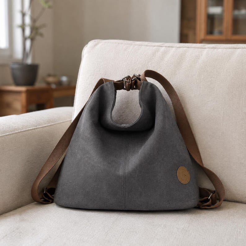 Puede incluir: Una mochila de lona gris oscuro con una silueta holgada y sin estructura. Cuenta con correas de tela marr&oacute;n que se conectan en la parte superior con herrajes met&aacute;licos y un peque&ntilde;o parche circular de aspecto de cuero marr&oacute;n en la parte inferior derecha.