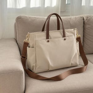 Könnte beinhalten: Eine beige Tragetasche mit dunkelbraunen Griffen und einem passenden verstellbaren Schultergurt. Die Tasche verfügt über eine Außentasche vorne mit zwei Druckknöpfen, Seitentaschen und goldfarbene Metallbeschläge für den abnehmbaren Gurt.