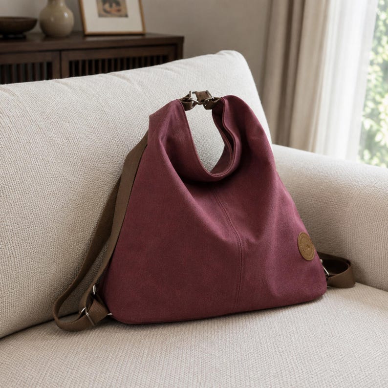 Puede incluir: Una mochila de lona color burdeos intenso con silueta triangular, correas ajustables marrones y un parche circular marr&oacute;n en la parte inferior derecha. El bolso presenta una costura vertical central y herrajes met&aacute;licos en la parte superior.