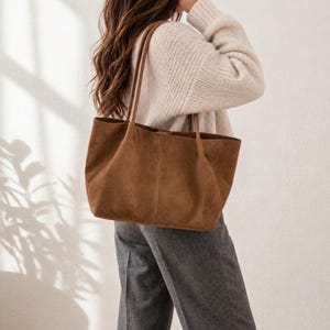 May include: A large, unstructured brown suede tote bag with two long shoulder straps. The bag features a simple, open-top design with a vertical seam running down the center of the front panel, providing a spacious and functional carryall accessory.