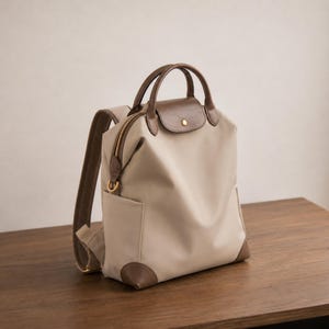 Op de afbeelding: Een beige rugzak met bruine lederlook afwerking, voorzien van een handvat aan de bovenkant, een overslagflap met een goudkleurige drukknoopsluiting, een hoofdvak met ritssluiting, zijvakken en verstelbare schouderbanden met een mesh achterpaneel.
