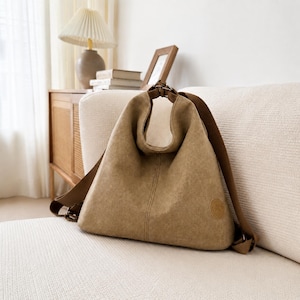 Puede incluir: Un bolso hobo de lona color canela con una silueta triangular holgada, que presenta una correa de hombro integrada, una costura vertical central y un peque&ntilde;o parche circular en la esquina inferior derecha. El bolso tiene herrajes met&aacute;licos.