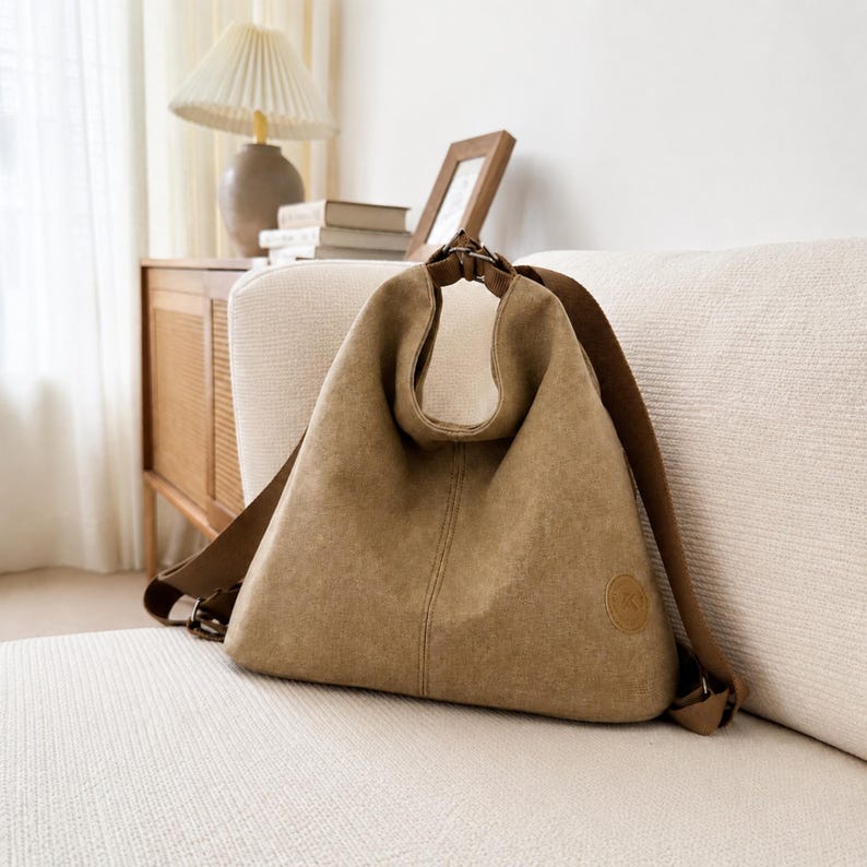 Puede incluir: Un bolso hobo de lona color canela con una silueta triangular holgada, que presenta una correa de hombro integrada, una costura vertical central y un peque&ntilde;o parche circular en la esquina inferior derecha. El bolso tiene herrajes met&aacute;licos.