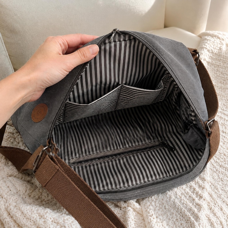 Puede incluir: Un bolso bandolera de lona gris carb&oacute;n con correa ajustable marr&oacute;n y cierre de cremallera. El interior est&aacute; forrado con tela de rayas verticales blancas y negras, con dos bolsillos abiertos y un bolsillo horizontal con cremallera.