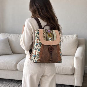 May include: A backpack featuring a cream-colored fabric with a multi-colored floral and bird pattern, a tan suede-like flap with a decorative woven strip, a brown faux leather lower section with fringe accents, and a large wooden button closure.