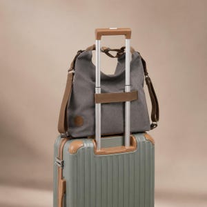 Puede incluir: Un bolso tote gris carb&oacute;n con correas marrones y un parche circular marr&oacute;n, que cuenta con una cinta horizontal en la parte posterior para sujetarlo al equipaje. El bolso est&aacute; colocado sobre el asa de una maleta verde salvia con esquinas.