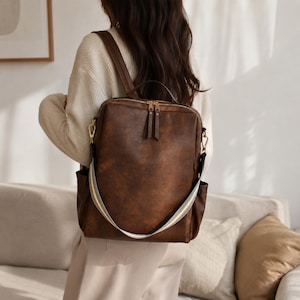 Peut inclure: Un sac à dos rectangulaire marron foncé à la finition lisse, doté de fermetures éclair en métal doré, d'une poignée supérieure et d'une bandoulière amovible à rayures multicolores blanches, jaunes et bleues. Le sac possède des poches latérales.