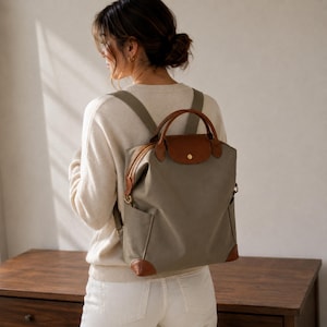 May include: A sage green backpack with brown leather accents, featuring a top handle, a fold-over flap with a snap closure, adjustable shoulder straps, and side pockets. The bag has a structured design with reinforced leather corners at the base.