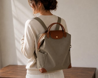 Puede incluir: Una mochila de color verde salvia con detalles en cuero marrón, que cuenta con un asa superior, una solapa con cierre de broche, correas ajustables para los hombros y bolsillos laterales. El diseño es estructurado con esquinas de cuero.