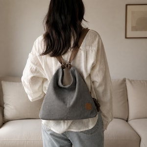 Puede incluir: Una mochila de lona gris con una silueta holgada y triangular y correas marrones. El bolso presenta un parche circular marrón en la parte inferior derecha y herrajes metálicos que conectan las correas con la abertura del compartimento.