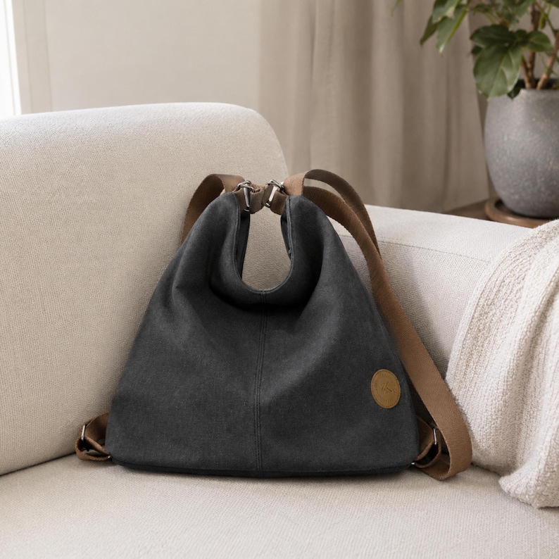 Puede incluir: Una mochila de lona gris carb&oacute;n con una silueta holgada y sin estructura. Cuenta con correas de hombro anchas de tela color canela, herrajes de metal plateado y un peque&ntilde;o parche circular de cuero sint&eacute;tico color canela en la esquina inferior derecha.