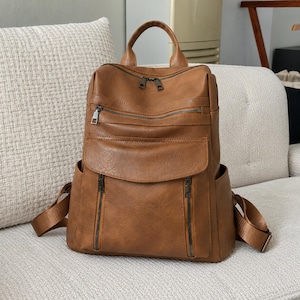 May include: A tan brown backpack featuring a top handle, adjustable shoulder straps, and multiple exterior compartments. The bag includes a main zippered section, a horizontal zippered pocket, a front flap pocket, and two vertical zippered pockets.