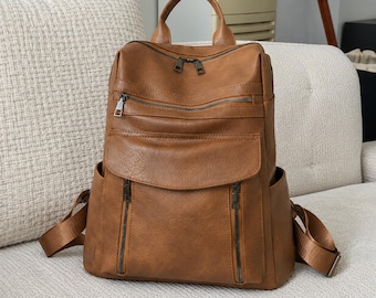 Puede incluir: Una mochila de color marrón claro con asa superior, correas ajustables para los hombros y múltiples compartimentos exteriores. La bolsa incluye una sección principal con cremallera, un bolsillo horizontal, un bolsillo frontal con solapa y dos verticales.
