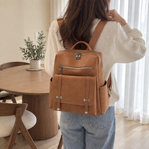 May include: A tan backpack with a smooth texture, featuring a top handle, adjustable shoulder straps, a zippered main compartment, a horizontal zippered front pocket, and a large flap-covered front pocket with two vertical decorative straps.