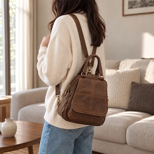 Puede incluir: Una mochila marrón con acabado texturizado, que cuenta con un asa superior, correas ajustables para los hombros y un bolsillo frontal con solapa y detalles de costuras. El bolso incluye herrajes metálicos, como cremalleras y anillas.