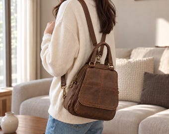 Puede incluir: Una mochila marrón con acabado texturizado, que cuenta con un asa superior, correas ajustables para los hombros y un bolsillo frontal con solapa y detalles de costuras. El bolso incluye herrajes metálicos, como cremalleras y anillas.
