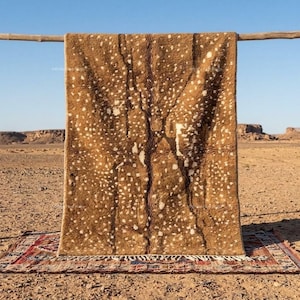May include: A rectangular rug with a deep brown base color featuring a dense pattern of small, irregular white spots across the surface. The rug has a thick, textured pile and a fringed edge at the bottom, hanging from a horizontal wooden pole.
