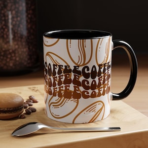 Peut inclure: Un mug en céramique blanche avec un intérieur et une anse noirs, orné d'un motif répétitif de contours de grains de café marron et du mot COFFEE imprimé dans une police marron foncé ondulée et empilée sur la surface extérieure.
