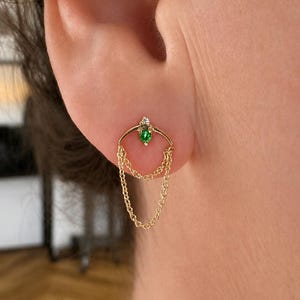以下が含まれることがあります： 小さな丸い緑色の宝石と上部に極小の透明なアクセントストーンをあしらった、曲線的なアーチデザインのゴールドピアス。アーチのベースから2本の繊細なゴールドチェーンがループ状に垂れ下がっています。