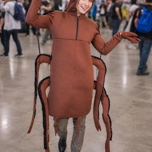 Pode incluir: Um traje de barata castanho composto por um top com capuz e antenas longas, fecho frontal e quatro pernas de inseto presas em cada lado. O traje é usado sobre calças cinzentas e botas pretas.
