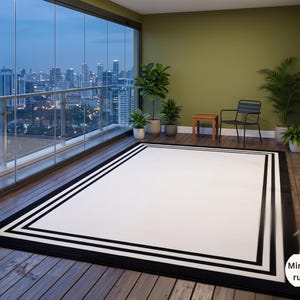 May include: A rectangular area rug featuring a large white center panel framed by multiple bold black and white border stripes. The rug is positioned on a wooden deck floor. A white logo with the text Minahil rugs is visible in the corner.