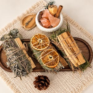 May include: A dark wooden tray holds three bundles of dried herbs and wood sticks tied with twine, decorated with two dried orange slices. A small white bowl contains pink salt chunks, a cinnamon stick, and a pinecone sits nearby.