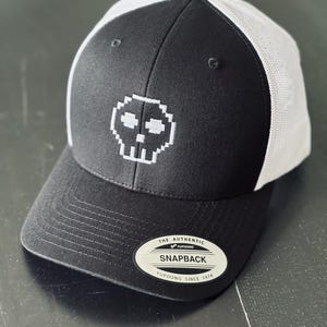 Op de afbeelding: Een zwart-witte truckerpet met een geborduurde, wit gepixelde schedel op het voorpaneel. De pet heeft een zwarte klep en voorkant, witte mesh achterpanelen en een ronde sticker op de klep met de tekst The Authentic Yupoong Snapback.