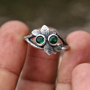 Op de afbeelding: Een zilveren ring met een textuur die lijkt op boomtakken, versierd met twee ronde, gefacetteerde groene stenen en gedetailleerde bladvormige accenten. De tekst TSHILVERGMES is zichtbaar op de ring. Het ontwerp bevat organische, botanische elementen.