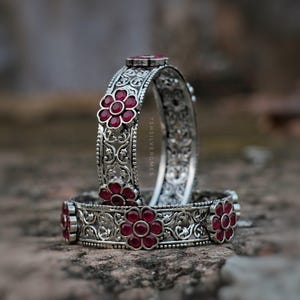 Könnte beinhalten: Zwei Armreifen aus silberfarbenem Metall mit komplexen Filigranmustern und eingelassenen dunkelroten blumenförmigen Steinen. Der Text TSHILVERGMES ist vertikal in die Seite des aufrechten Armreifs eingraviert und zeigt ein detailliertes Metalldesign.