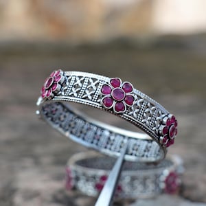 Könnte beinhalten: Ein Armreif aus oxidiertem Silber mit einem sich wiederholenden Muster aus erhabenen Metallblumenmotiven, die jeweils mit kleinen runden rosa Steinen besetzt sind. Das Band hat ein komplexes Gitterdesign und der Text TSHSILVERONES ist sichtbar.