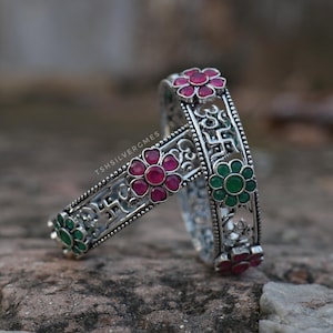 Könnte beinhalten: Zwei silberne Armreifen mit oxidierten Metallarbeiten und abwechselnd rosa und grün geformten Blumensteinen. Das Design enthält geprägte Om- und Swastika-Symbole entlang des Bandes. Der Text TSHSILVERGMES ist auf der Metalloberfläche sichtbar.