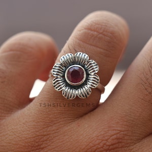 Könnte beinhalten: Ein Silberring mit einer blumenförmigen Fassung und einem runden, facettierten dunkelroten Edelstein in der Mitte. Das Metall hat eine texturierte, oxidierte Oberfläche mit sichtbaren Rillen auf den Blütenblättern. Der Text TSHSILVERGEMS ist abgebildet.