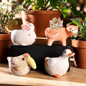 Puede incluir: Cuatro macetas de cerámica con forma de animal, incluyendo un conejo blanco, un zorro color melocotón con una suculenta, un pájaro moteado con pico amarillo y un pato blanco con detalles naranjas, sobre un bloque rectangular negro.