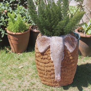 May include: A cylindrical brown planter with a textured surface featuring a sculpted elephant head on the front. The elephant has large ears and a trunk extending downward. A vibrant green fern plant is growing out of the top of the planter.