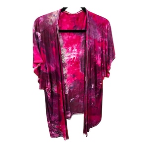 Puede incluir: Un kimono de color rosa y morado con estampado tie-dye y mangas cortas. El kimono está hecho de una tela suave y fluida y tiene un corte holgado.