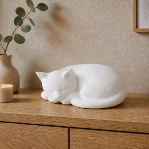 Könnte beinhalten: Eine mattweiße Skulptur einer schlafenden Katze, die zu einer kompakten, abgerundeten Form zusammengerollt ist. Die Figur weist glatte, klare Linien mit subtilen Details an den Ohren, den geschlossenen Augen und den eingezogenen Pfoten auf.
