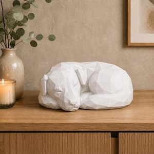 Op de afbeelding: Een wit, geometrisch low-poly beeldhouwwerk van een slapende hond. Het figuur is gemaakt met scherpe, hoekige facetten die de vorm van het rustende dier definiëren, dat opgerold ligt met de kop naar beneden. Het oppervlak is glad en mat.