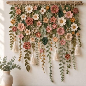 May include: A wall hanging featuring crocheted flowers and leaves in shades of pink, white, green, and yellow, suspended from a horizontal piece of driftwood. The arrangement includes various floral patterns, trailing vines, and four cream-colored tassels.