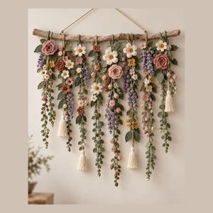 Può includere: Una decorazione da parete composta da un pezzo orizzontale di legno galleggiante con diversi fili di fiori e foglie all'uncinetto nei toni del rosa, bianco, giallo, viola e verde. La composizione include diverse nappe in filato color crema.
