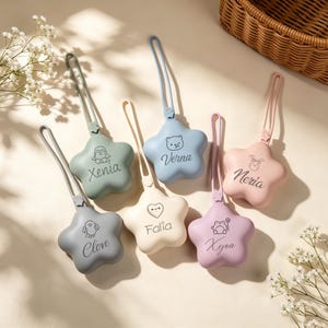 Peut inclure: Six étuis en silicone en forme d'étoile avec des boucles assorties, déclinés en sauge, bleu, crème, rose, gris et lavande. Chaque étui présente un personnage au trait et un nom : Clove, Xenia, Verna, Falia, Neria et Xyra, imprimés en gris foncé.