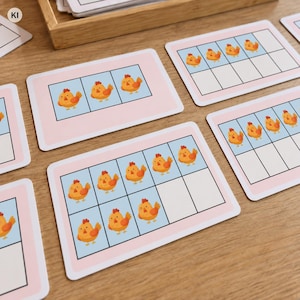 Op de afbeelding: Een set rechthoekige educatieve kaarten met afgeronde hoeken, elk voorzien van een lichtroze rand en een raster met getekende oranje kippen. De kaarten tonen variërende aantallen kippen in de rastervakken op een lichtblauwe achtergrond.