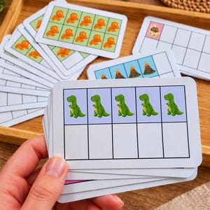 Puede incluir: Un mazo de tarjetas educativas rectangulares de diez marcos con ilustraciones de dinosaurios y volcanes. La tarjeta superior muestra cinco dinosaurios verdes de dibujos animados en la fila superior de una cuadrícula, con cinco cuadrados vacíos debajo. Otras tarjetas muestran dinosaurios naranjas.