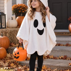 Puede incluir: Un poncho de fantasma en forro polar blanco con capucha que incluye ojos y boca de fieltro negro, combinado con leggings y botas negras. La prenda presenta una cara de fantasma ovalada en negro. Se sostiene un cubo de calabaza de plástico naranja.